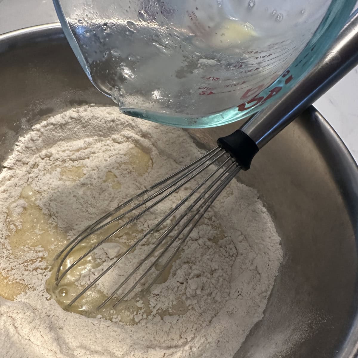 Wet ingredients poured from a glass bowl to whisked flour in a steel bowl.