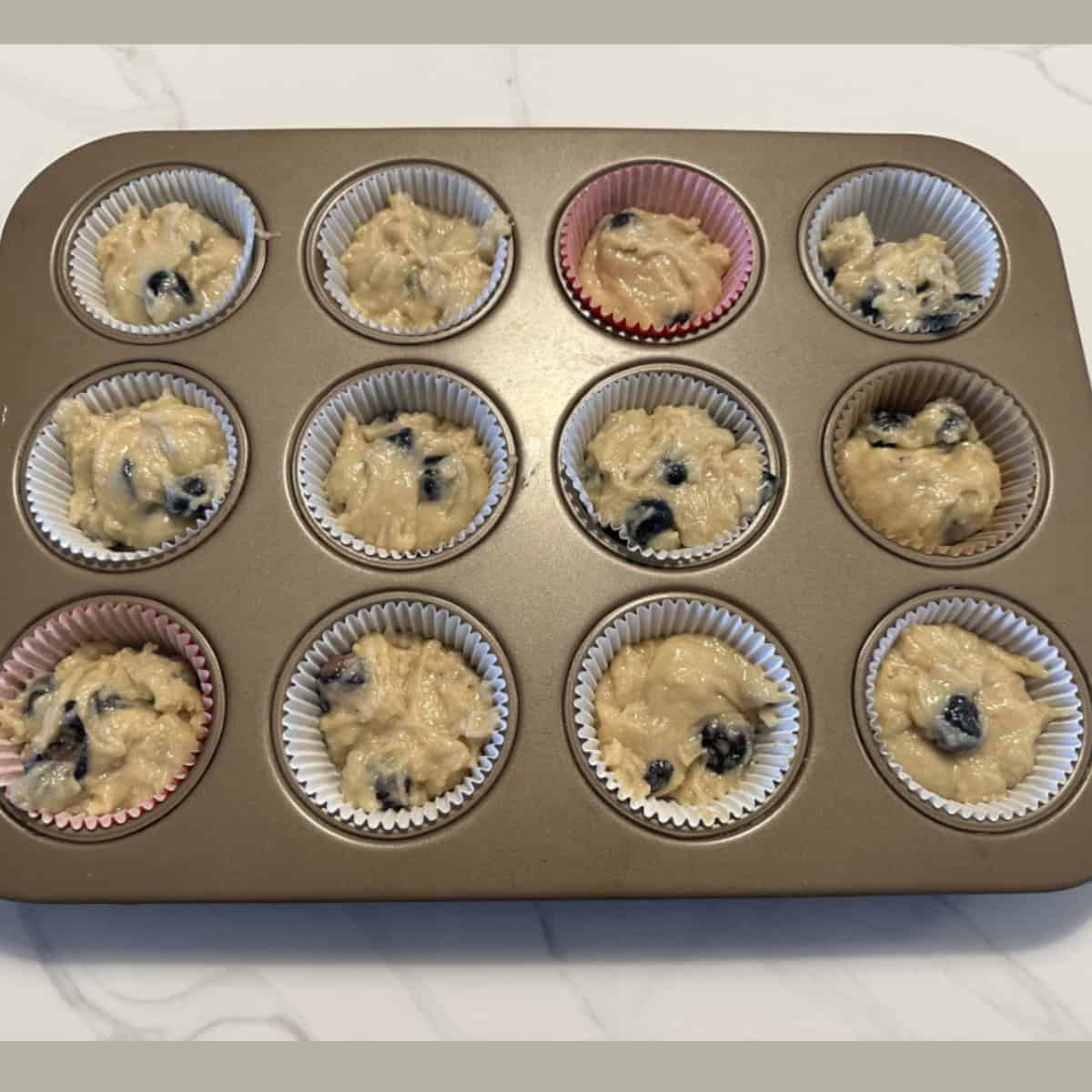 Blueberry muffin batter filled in paper cups in a muffin tray ready to bake.