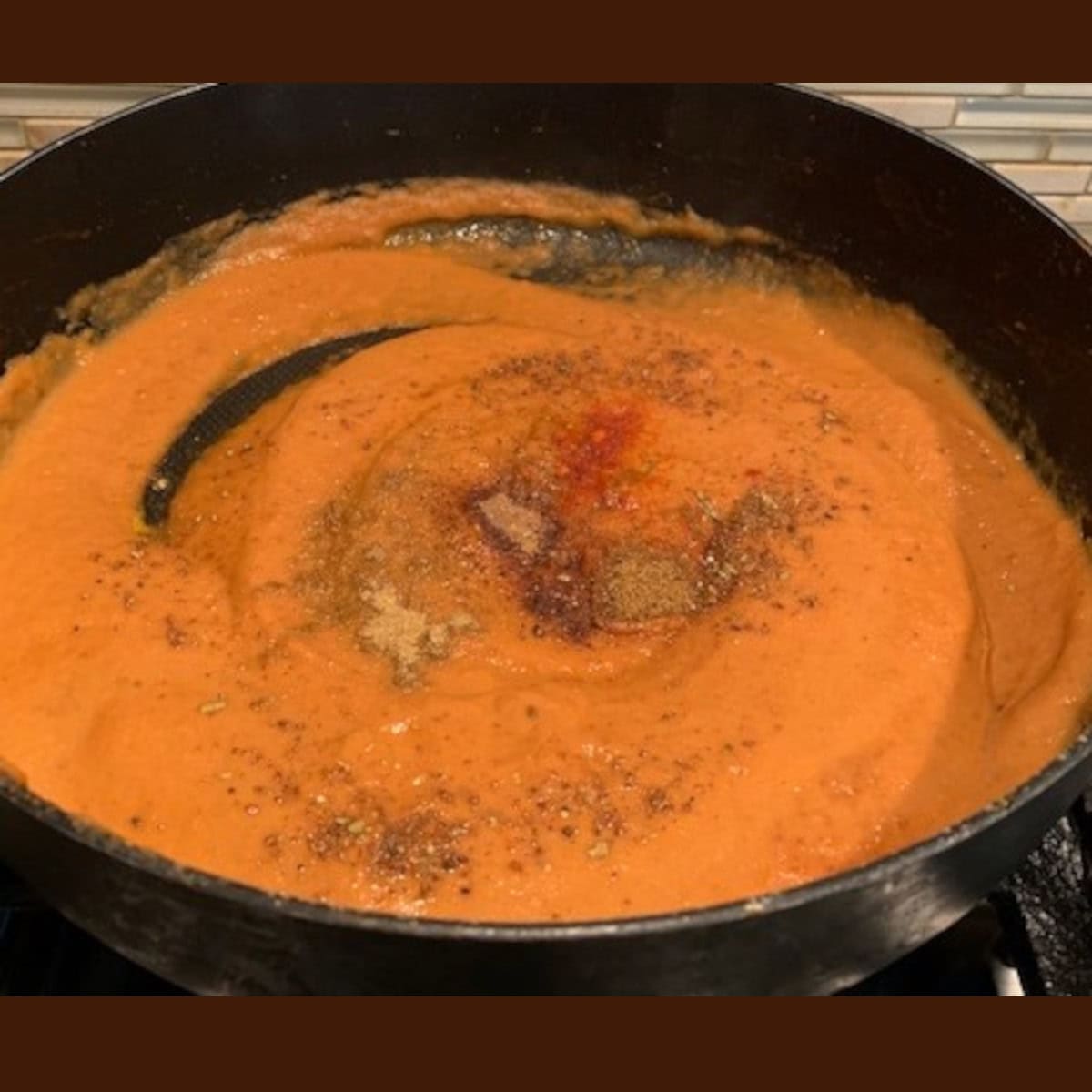 Spices added to chicken tikka masala gravy in a black pan on a stove top.