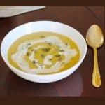 Roasted pumpkin soup made with Roasted pumpkin soup recipe in a white bowl , garnished with coconut milk and pumpkin seeds with a gold colored spoon next to it on a wooden table.