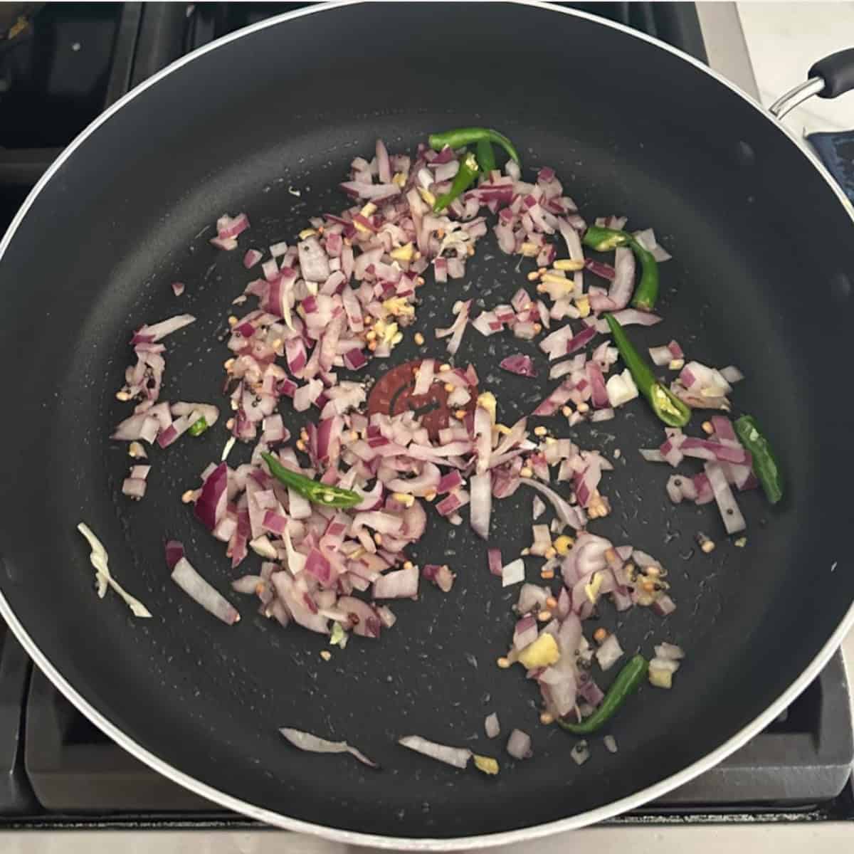 Cabbage coconut stir fry preparation with saute onions, green chilies and ginger in oil in a black pan on stove top.