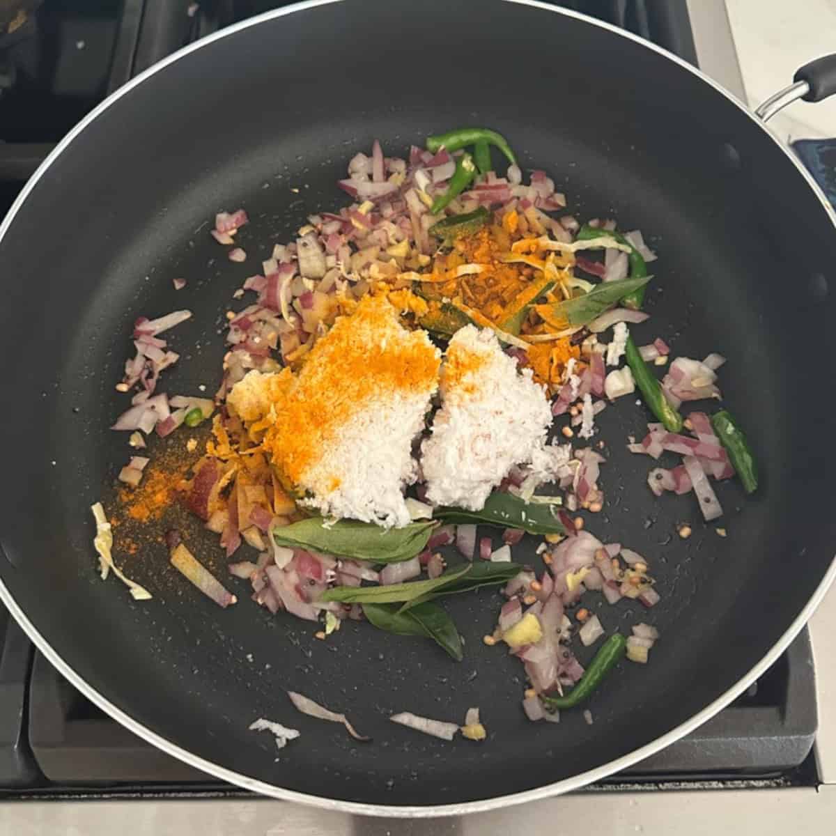Cabbage coconut stir fry preparation in a black pan on stove top with coconut , onions, green chilies.