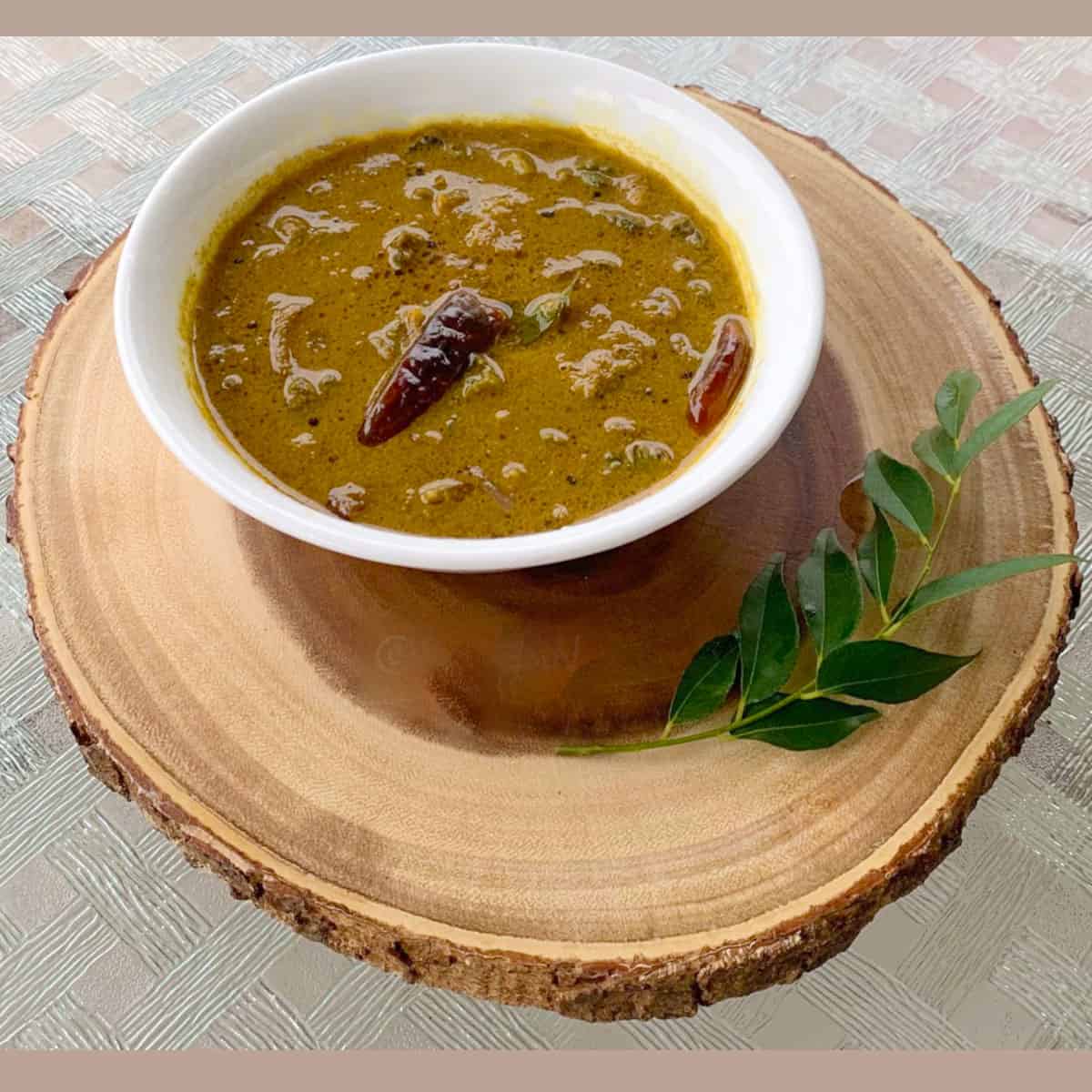 Pavakka Theeyal served in a white bowl on a wooden board with a curry leaf next to it.