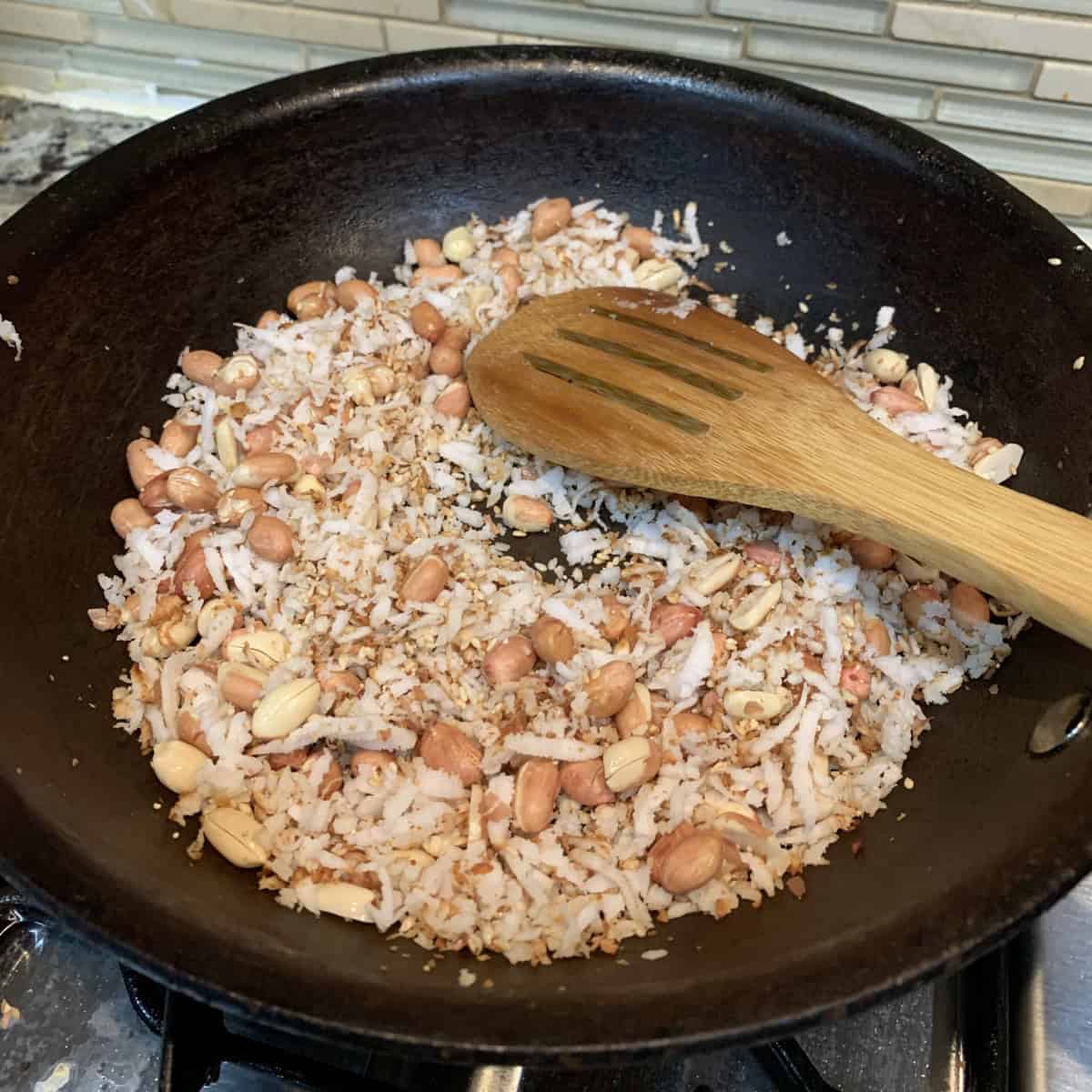 roasting coconut sesame and peanuts in a black pan with a wooden spoon.