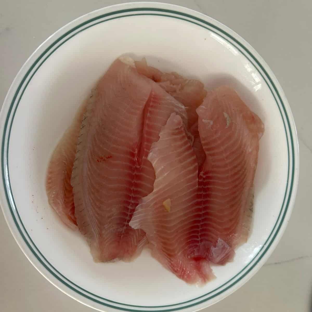 Tilapia fillets ready to be marinated in a white bowl.