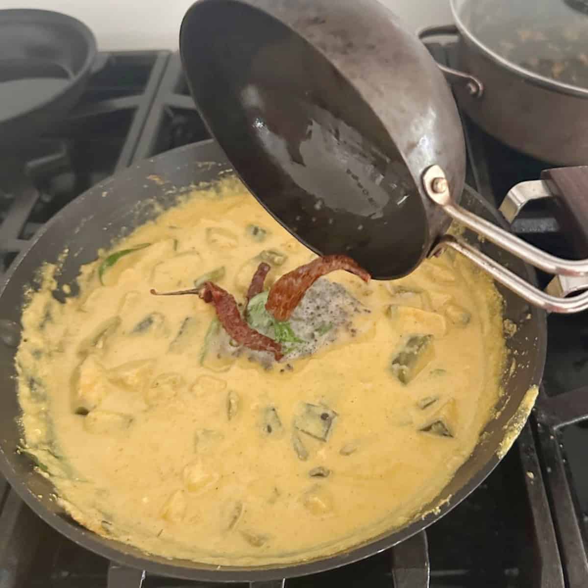 Tempering kaalan cooking in a black wok contents of tempering being poured from a small black pan.