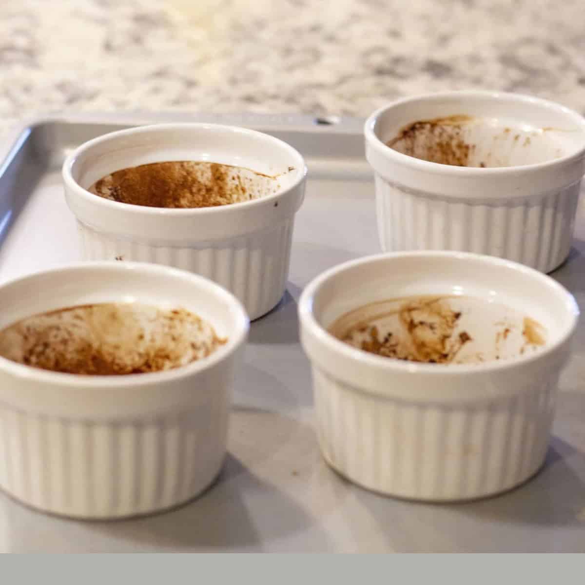 four white ramekins sprayed with oil and cocoa powder on a gray tray.