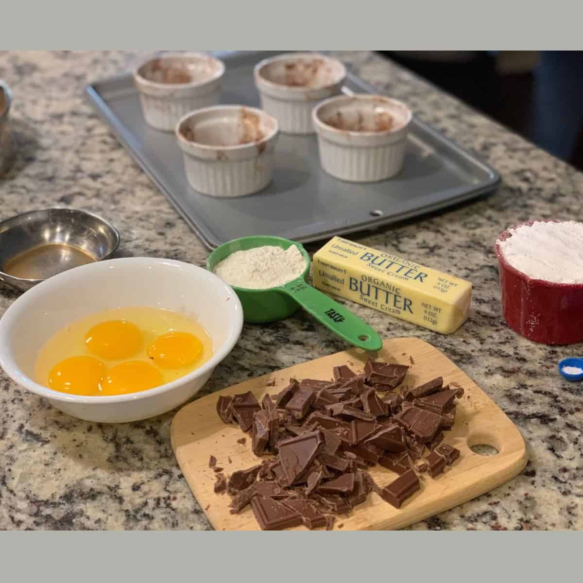 chocolate cut into pieces on a wooden board, eggs in a white bowl, along with other ingredients to make lava cake.