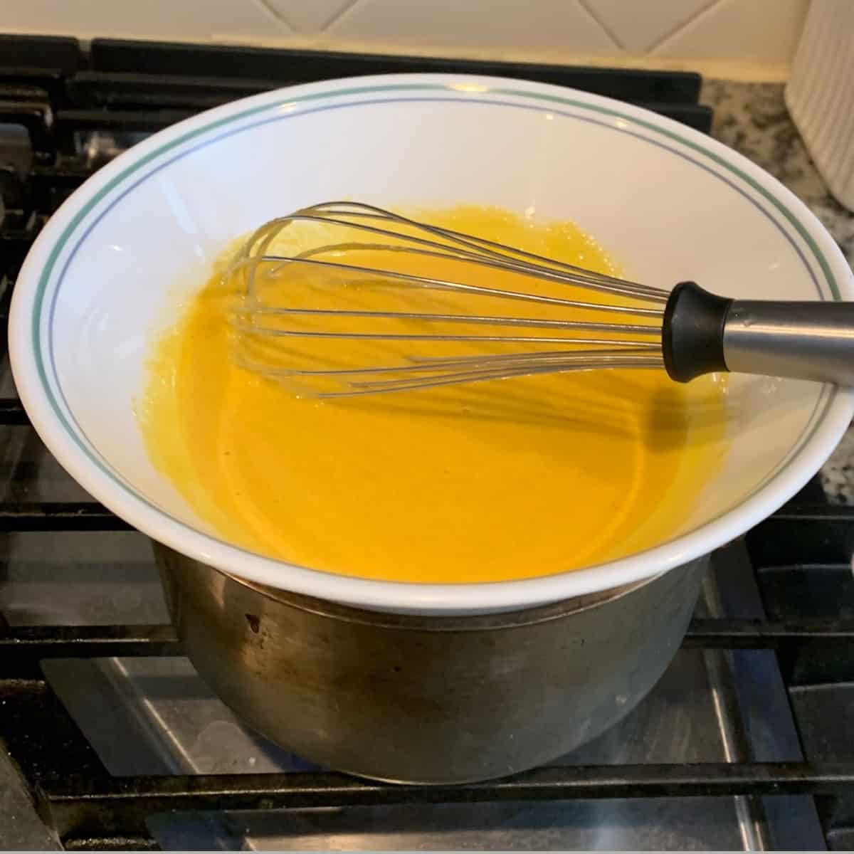 cooking eggs and sugar in a double boiler using a white bowl with eggs and whisk on top of steel container with boiling water on a gas stove .