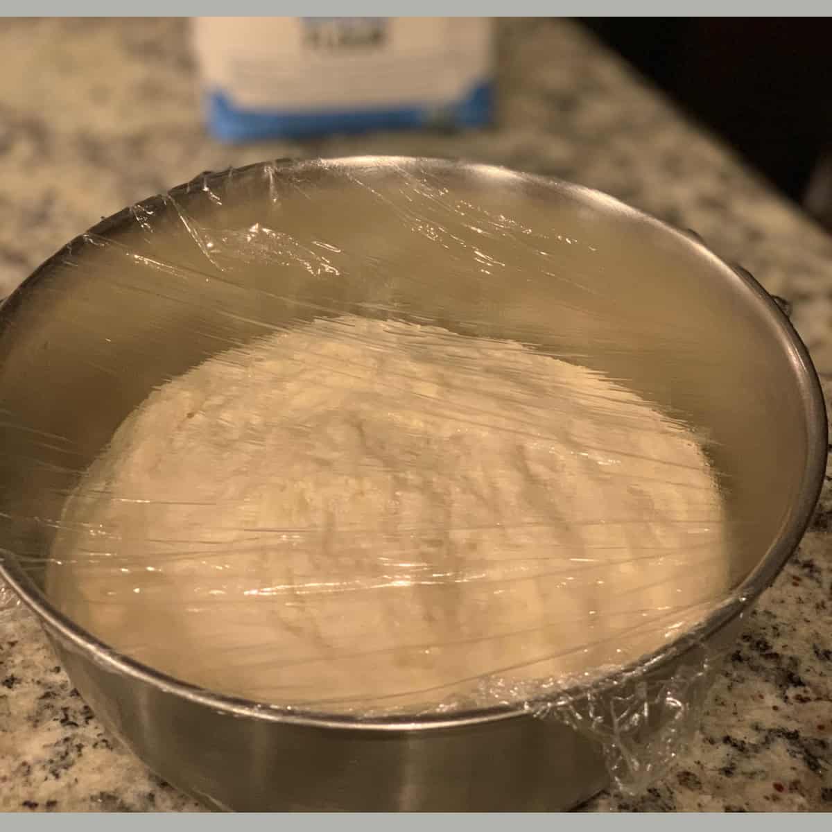 pizza dough doubled in size after fermentation in a silver bowl covered with a plastic wrap.
