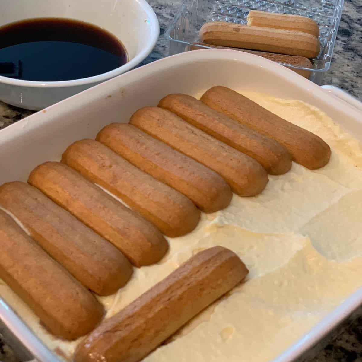 Layering the ladyfingers soaked in coffee to make Tiramisu in a white dessert bowl.