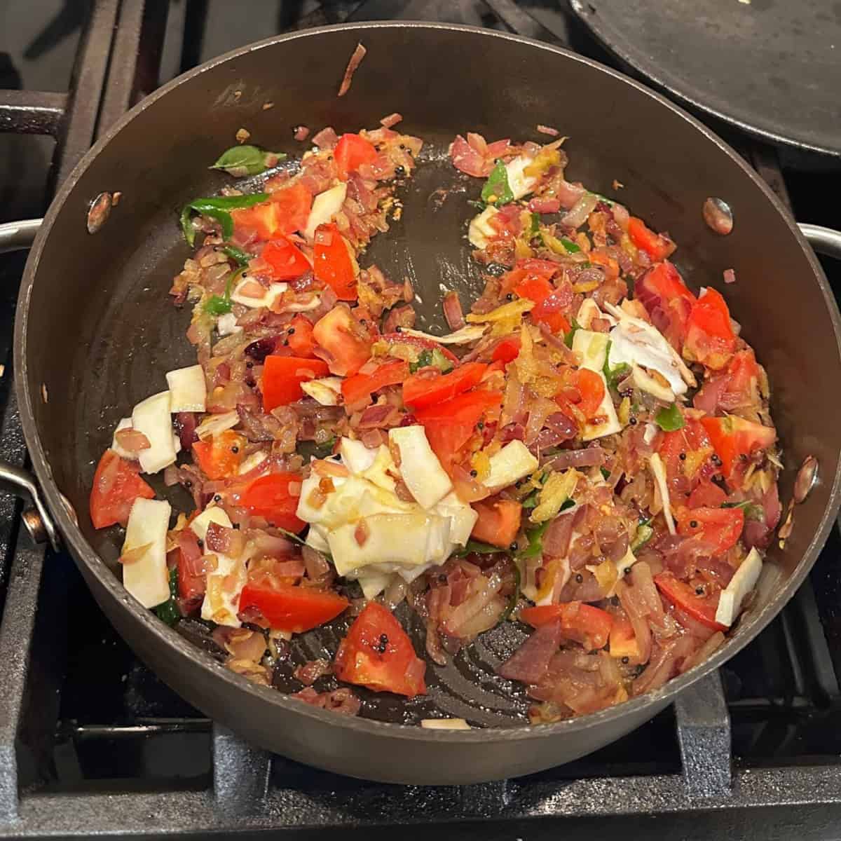 Tomatoes and coconut bits added to chemmeen roast gravy cooking in a black pan on a stove top.