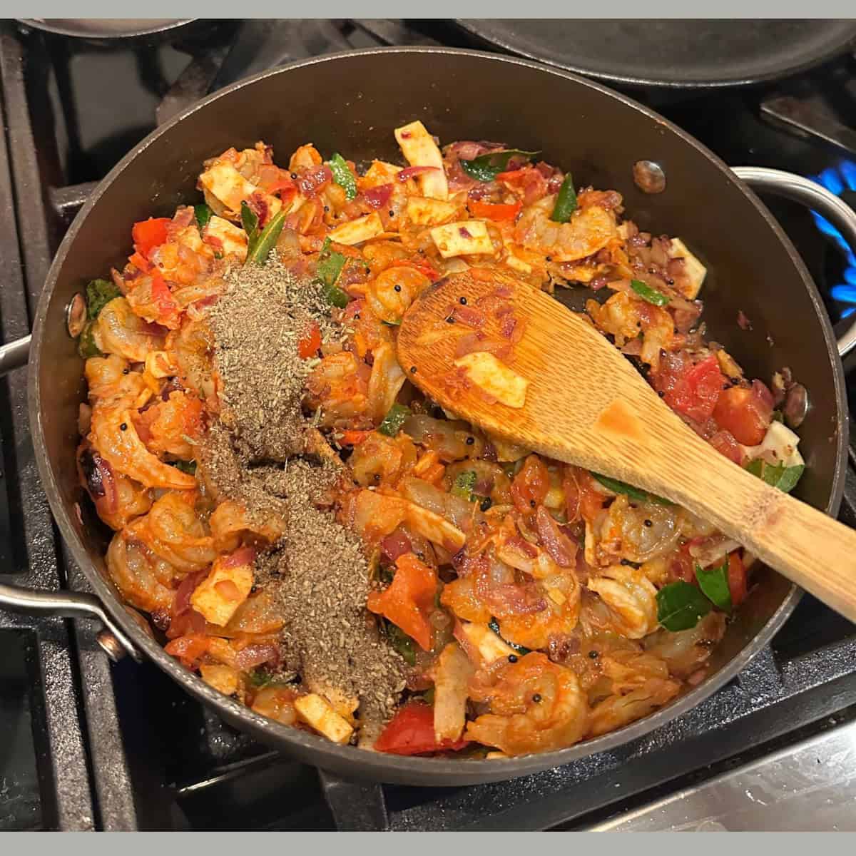 Spice powders being added to shrimp cooking in a pan with a wooden ladle being used to saute.