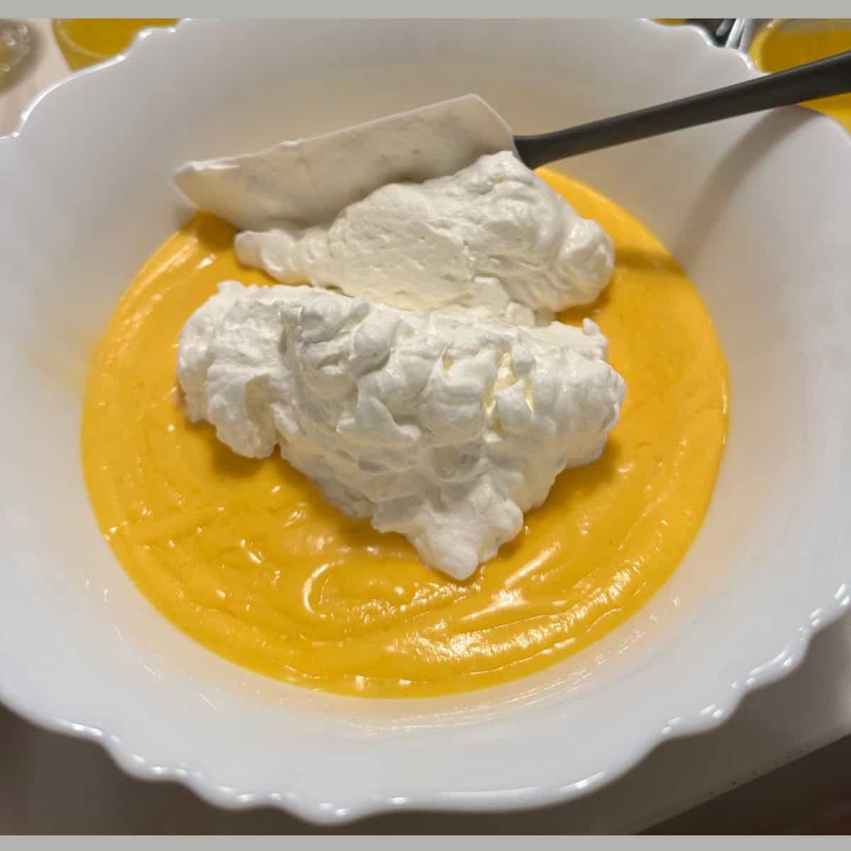 folding whipped cream and cooled mango gelatin mixture in a white bowl with a spatula.