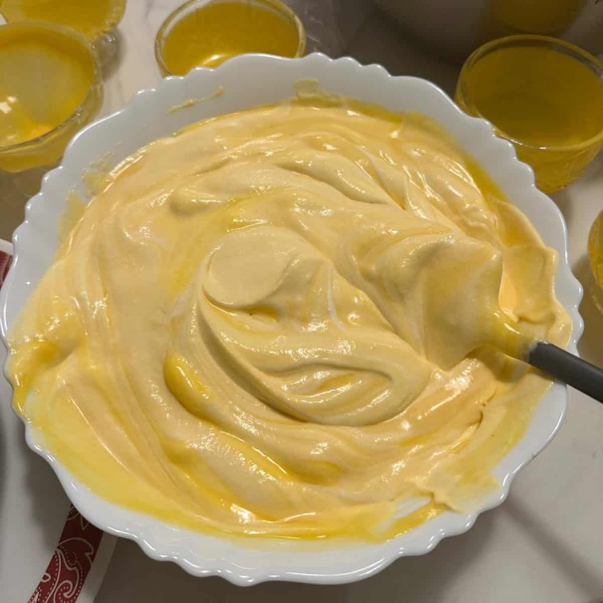 folding whipped cream and cooled mango gelatin mixture in a white bowl with a spatula.